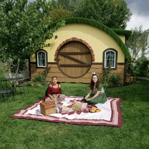 Two women sit on a picnic blanket in a garden, enjoying food in front of a whimsical, hobbit-style house.